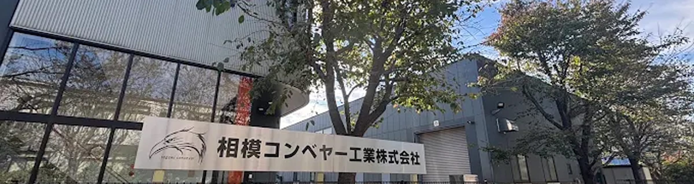 相模コンベヤー工業株式会社の社名看板と、木々に囲まれたオフィスビルディングの外観
