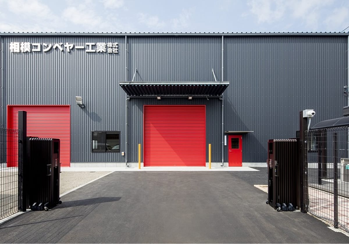 濃いグレーの外壁に赤いシャッターが映える、「相模コンベヤー工業株式会社」の社名が掲げられた工場の外観写真。