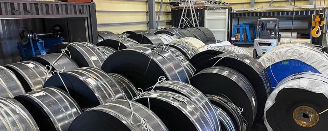 倉庫内に整然と保管されている、紐で固定された多数の黒いコンベアベルト用大型ロール