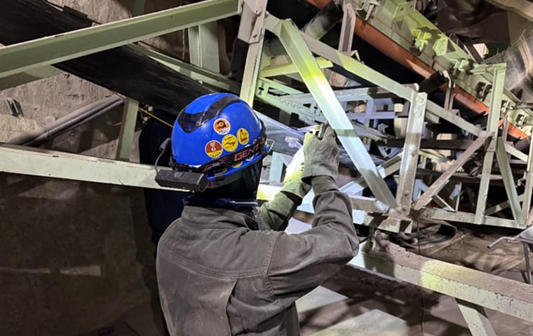 青いヘルメットを被った作業員が、暗所にあるベルトコンベアのフレームを点検している背後からのショット