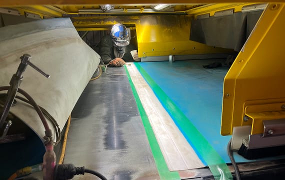 コンベア設備の内部で、ベルトの接合面や表面の調整作業を慎重に行うヘルメット姿の作業員