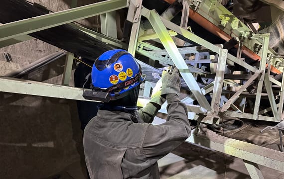 薄暗い屋内施設で、ステッカーの貼られた青いヘルメットを被った作業員が、ベルトコンベアのフレームやローラーの状態を間近で点検している様子。