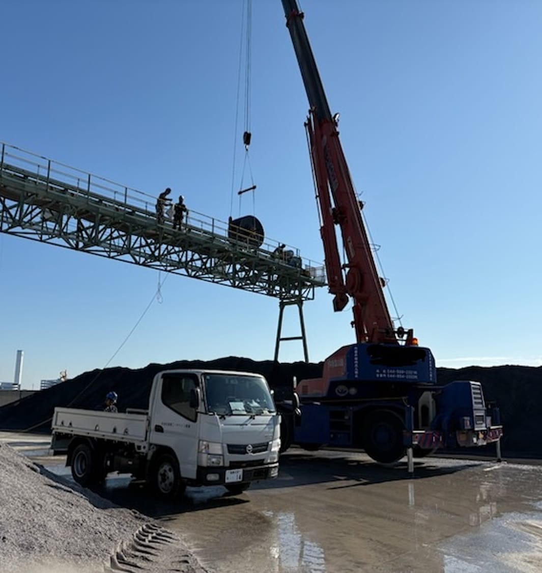 青空の下、大型の赤色クレーン車がベルトコンベアの設置ラインへ向かって、黒いゴムベルトのロールを吊り上げている屋外作業風景。