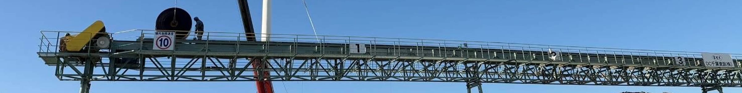 青空を背景に、トラス構造のフレームで支えられた非常に長い屋外ベルトコンベア。上部には点検用の通路と安全手すりが設置されている。