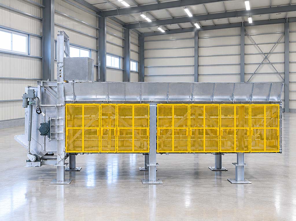工場内に設置された大型のエプロンコンベア。黄色の安全ガード付きで、金属製の構造が見える。.